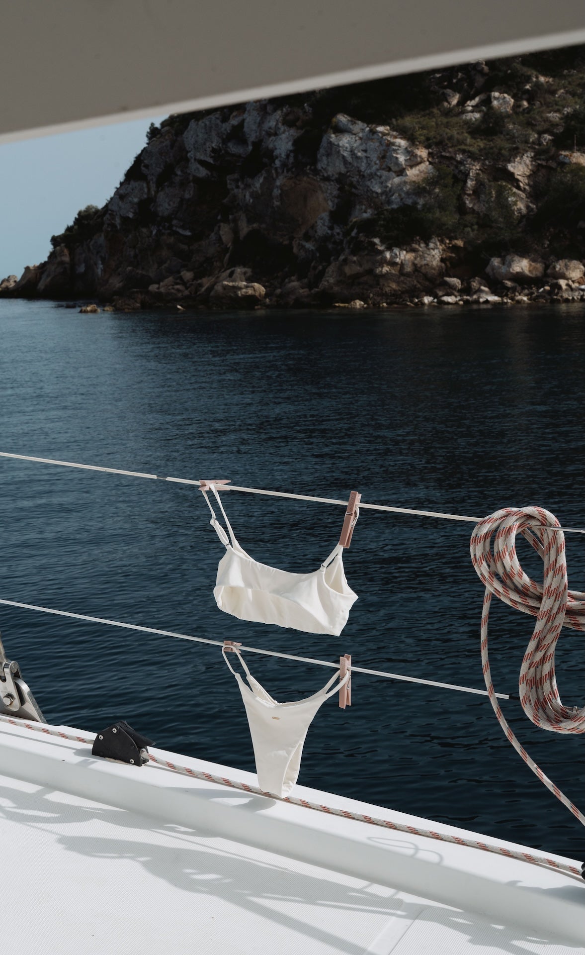 White bikini hanging on a clothesline with a scenic background of water and rocks.