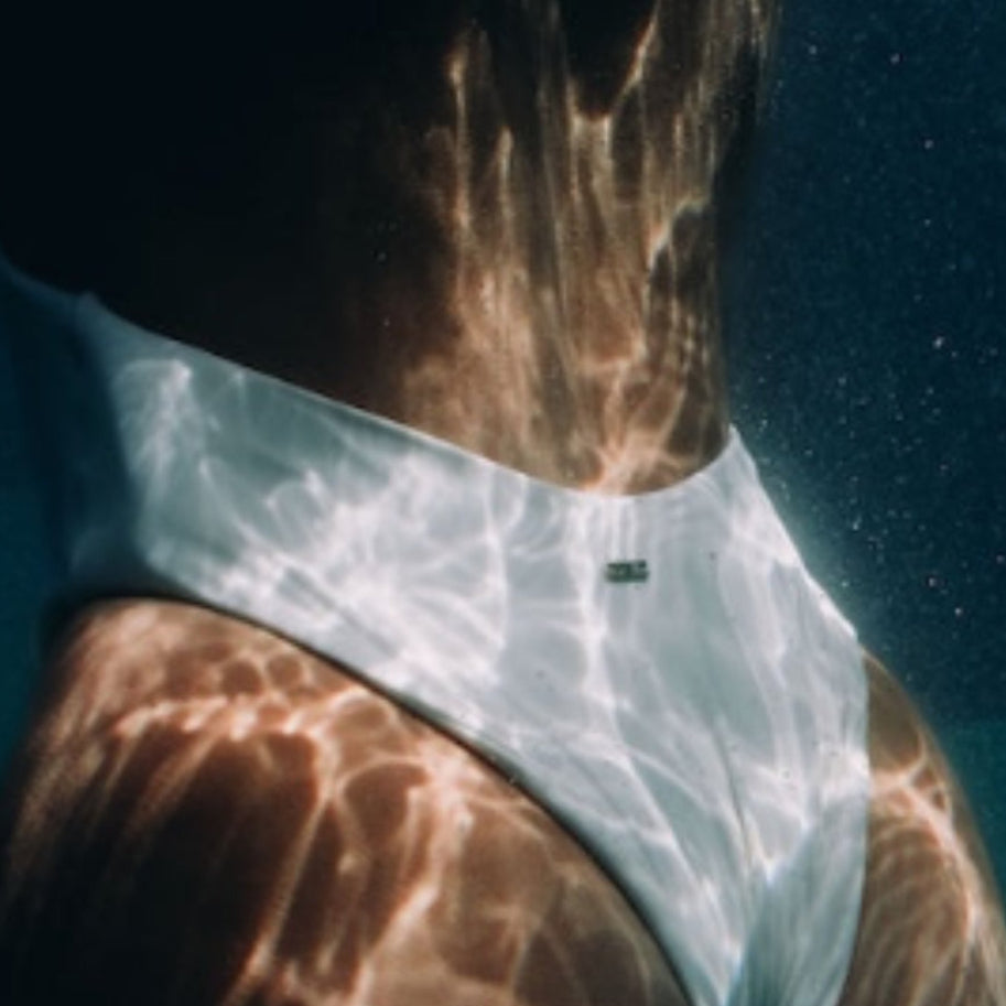 Person wearing a white swimsuit underwater with sunlight filtering through.