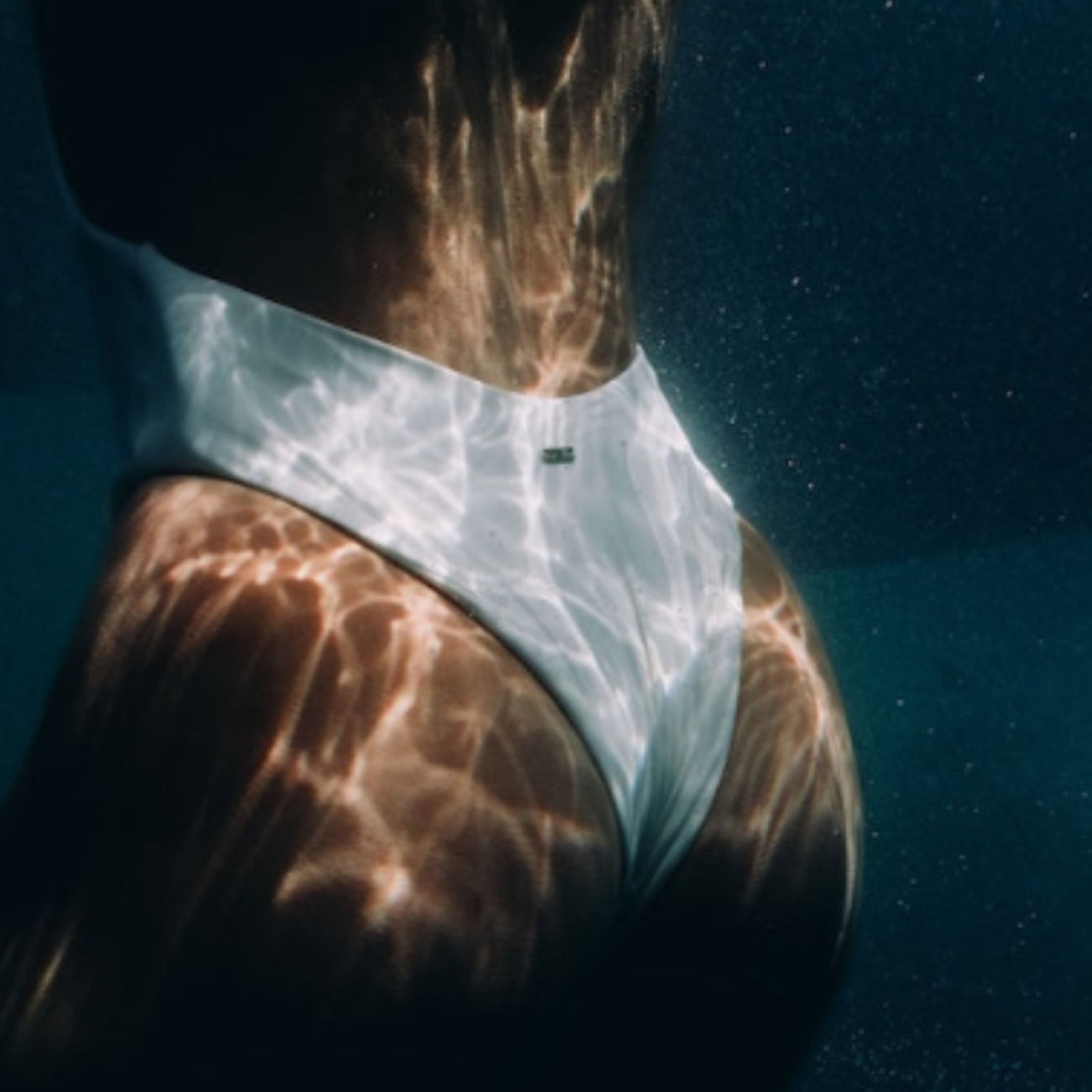 Person wearing a white swimsuit underwater with sunlight filtering through.